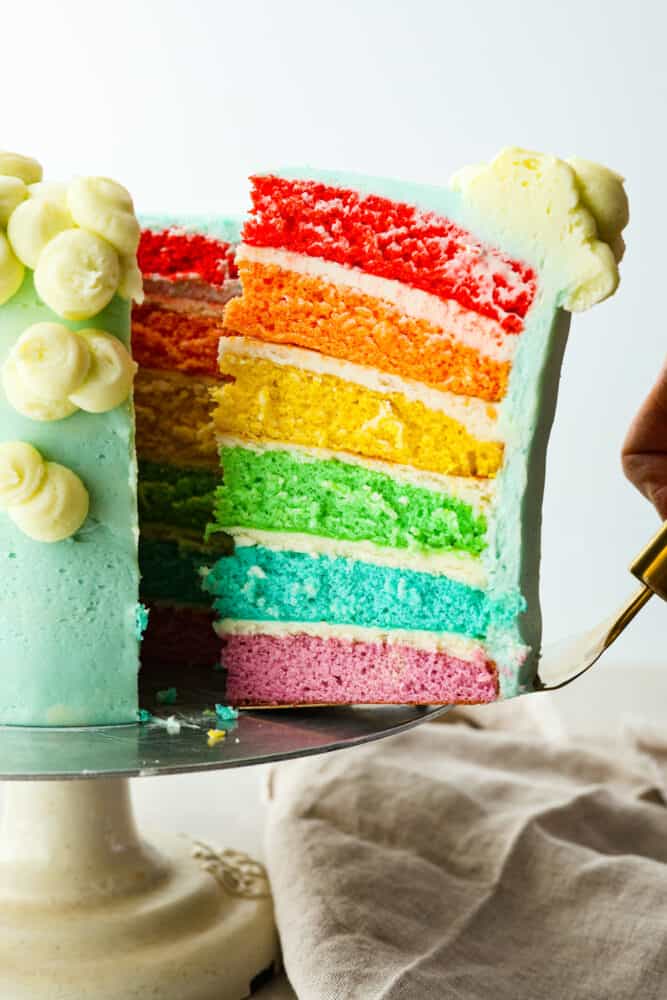 This gorgeous rainbow cake is the best way to add a splash of color to any special occasion! Not only is this cake beautiful to look at, but it’s extra moist and topped with luscious homemade buttercream. It’s the perfect dessert for birthdays, Saint Patrick’s Day, and more! Serving a piece of rainbow cake with a cake spatula.