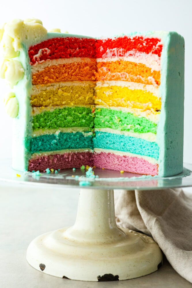 This gorgeous rainbow cake is the best way to add a splash of color to any special occasion! Not only is this cake beautiful to look at, but it’s extra moist and topped with luscious homemade buttercream. It’s the perfect dessert for birthdays, Saint Patrick’s Day, and more! A whole rainbow cake with a slice cut out of it.